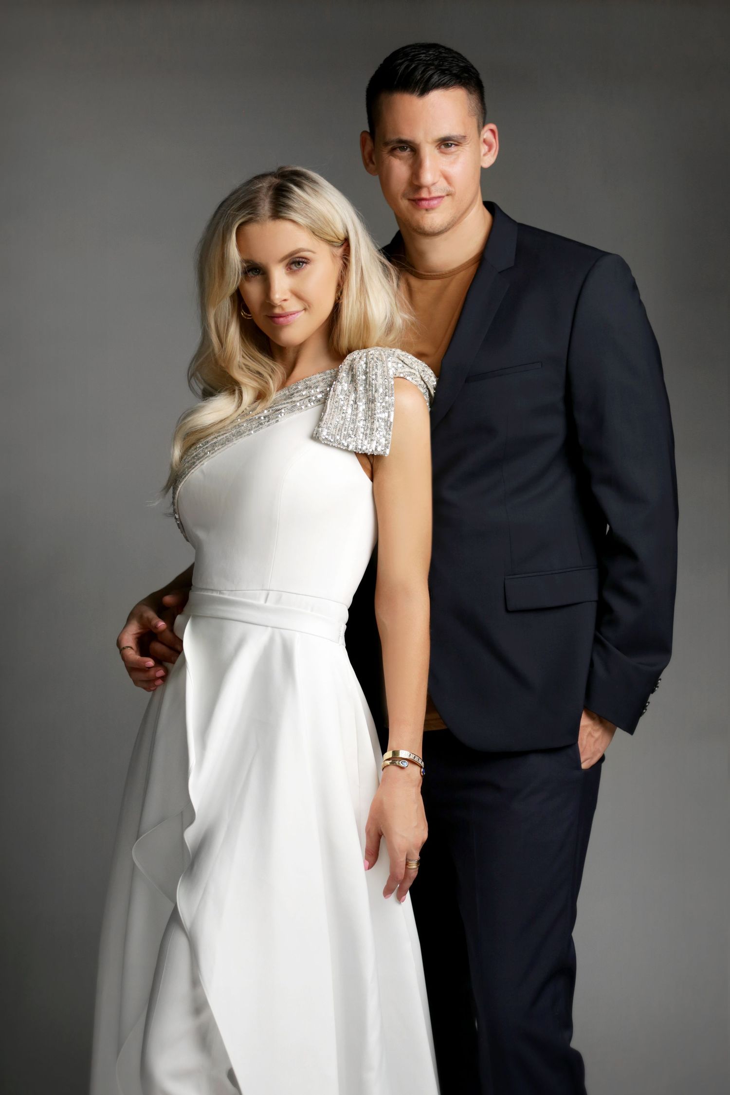 Elegant couple posing together, woman in white sequined gown and man in dark navy suit against gray background.