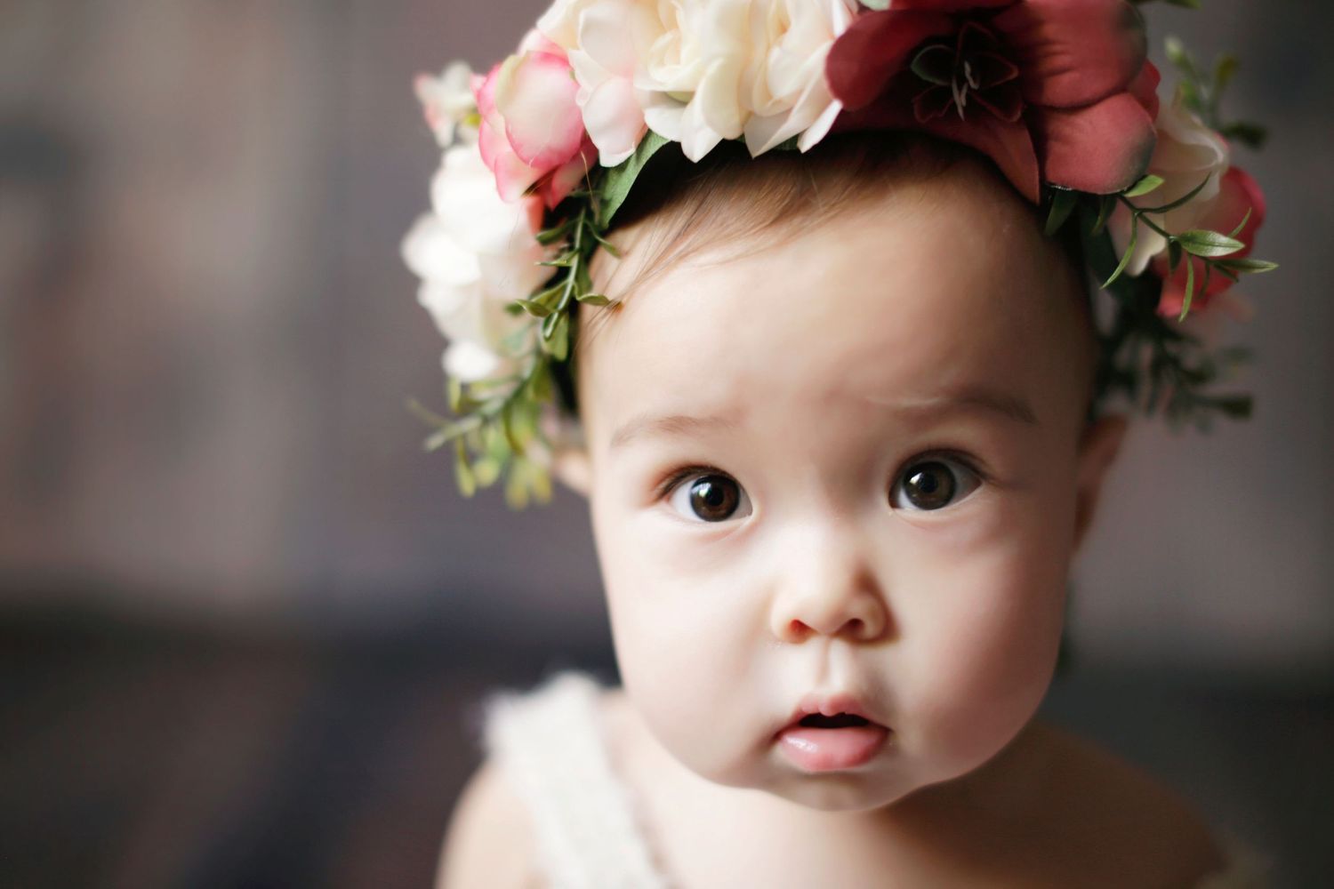 Adorable baby wearing a floral crown of pink and white flowers, looking curiously at the camera.