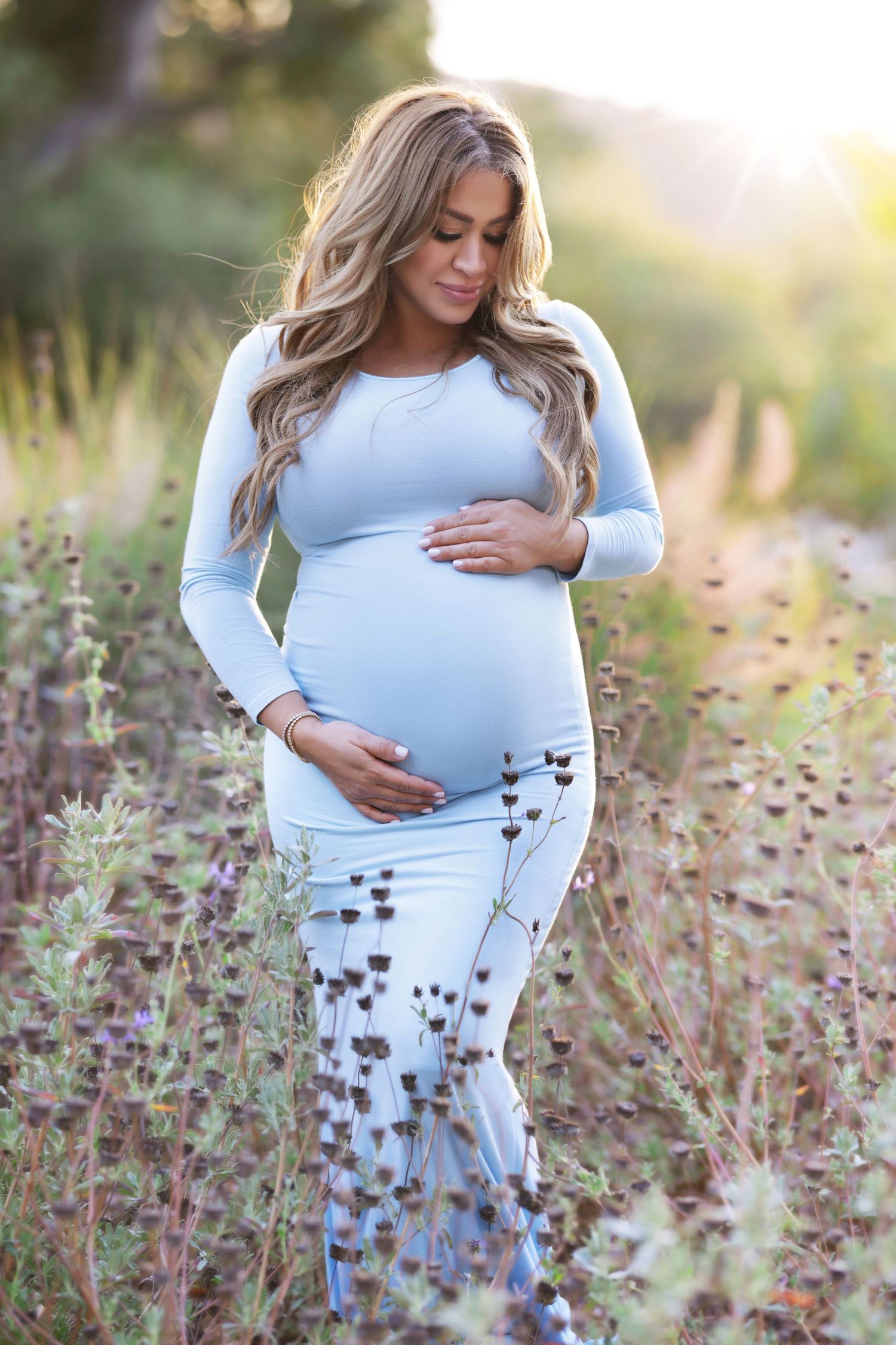 Pregnant woman in light blue dress cradling baby bump outdoors in a field of wildflowers during golden hour.