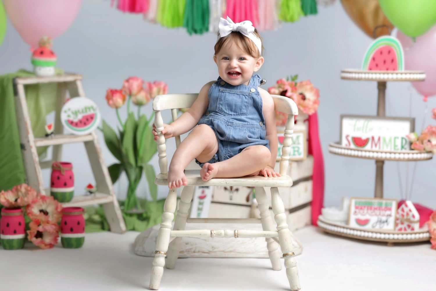 Cheerful toddler girl in denim overalls with white bow sitting on white bench with watermelon summer party decorations.
