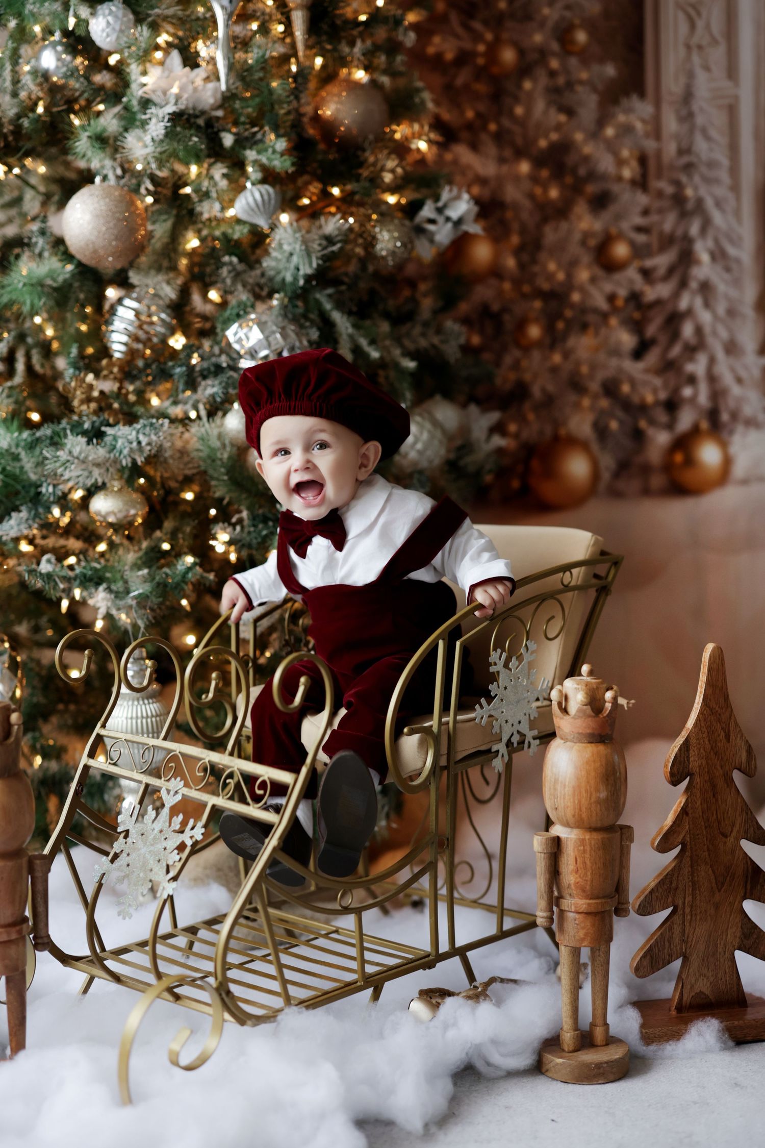 Happy baby in red velvet outfit sits in gold sleigh beside decorated Christmas tree with ornaments.