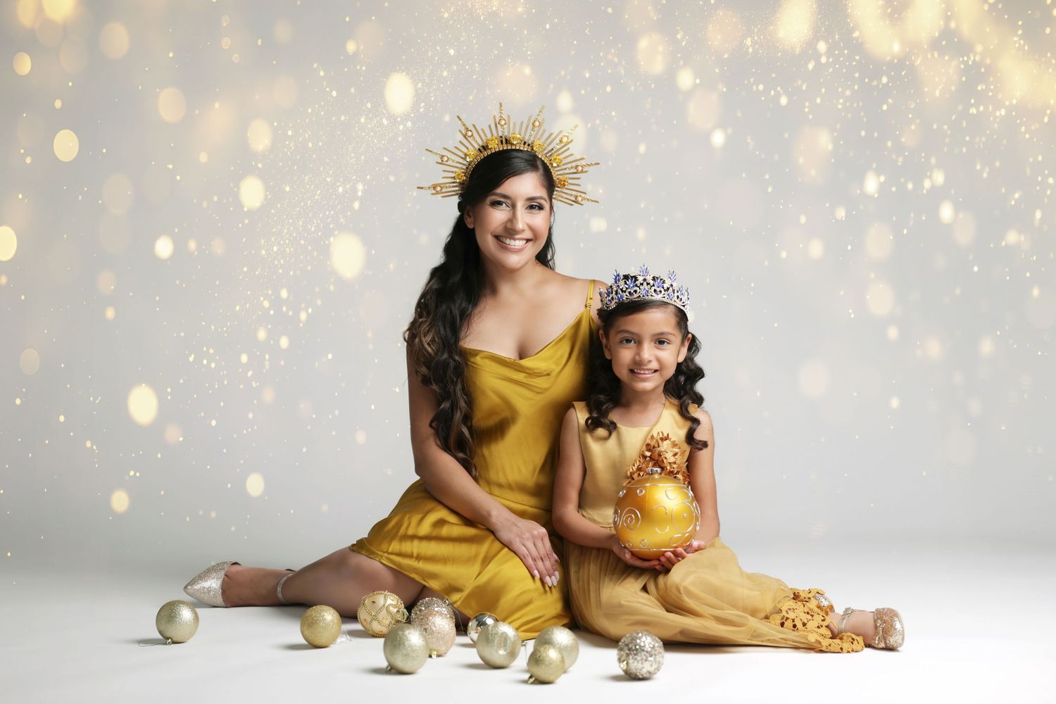 A woman and young girl in golden outfits pose with holiday ornaments against a sparkling bokeh background.