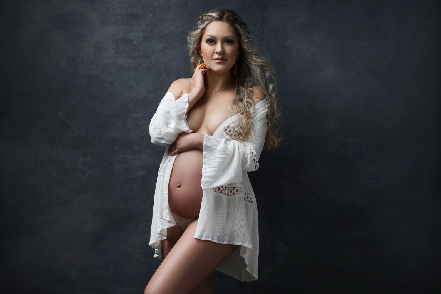 Pregnant woman in white lace robe posing for maternity photo shoot against dark background.
