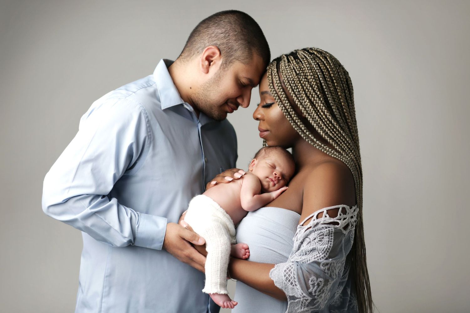 A couple tenderly holds their newborn baby in a professional studio portrait against a soft gray background.