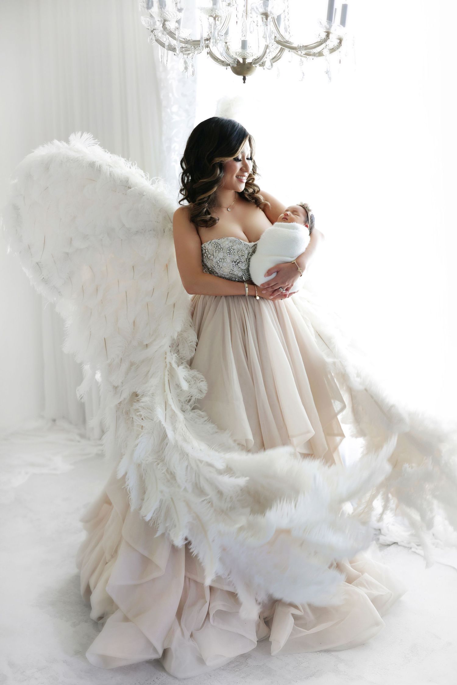Mother in angel wings and elegant gown tenderly holding newborn baby indoors.