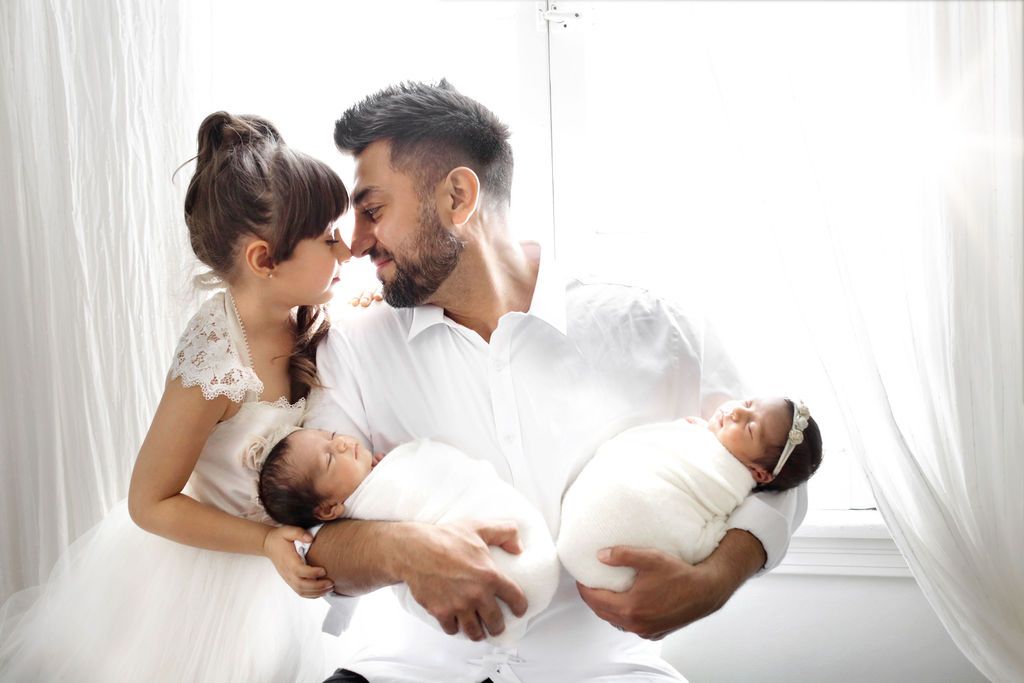 A father in white holds newborn twins while his young daughter in a white dress affectionately touches his face by a bright window.