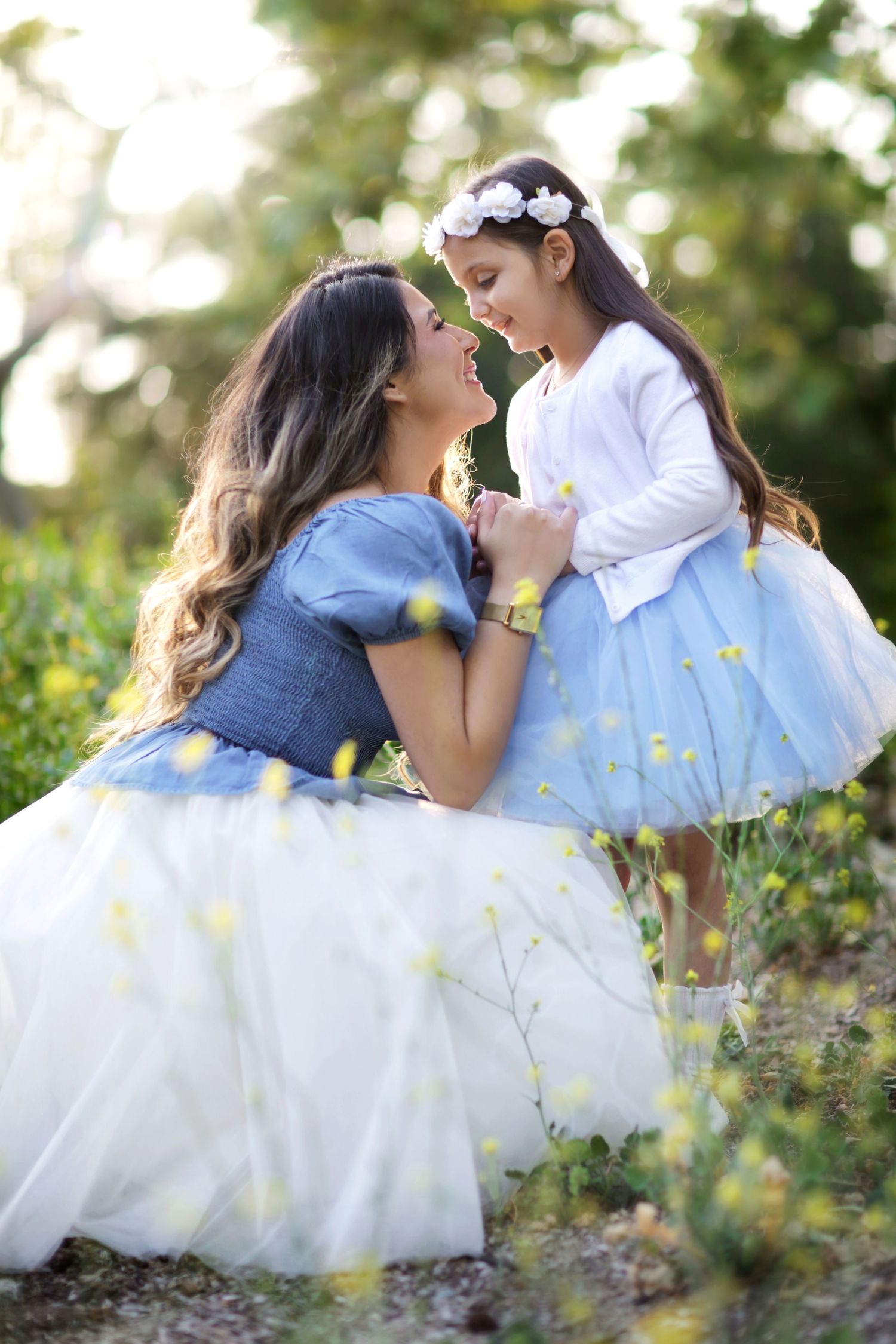 Mother and daughter in matching blue tulle dresses share a tender moment outdoors among yellow wildflowers.