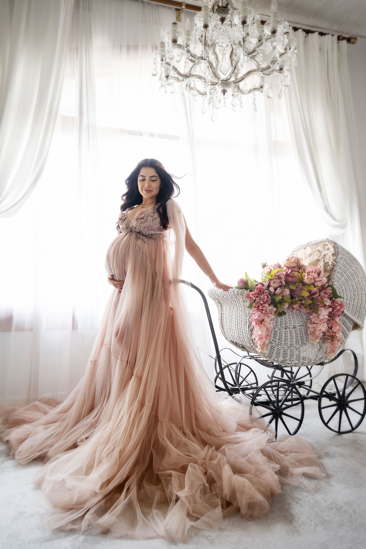 Pregnant woman in flowing blush tulle gown stands beside vintage floral baby carriage.