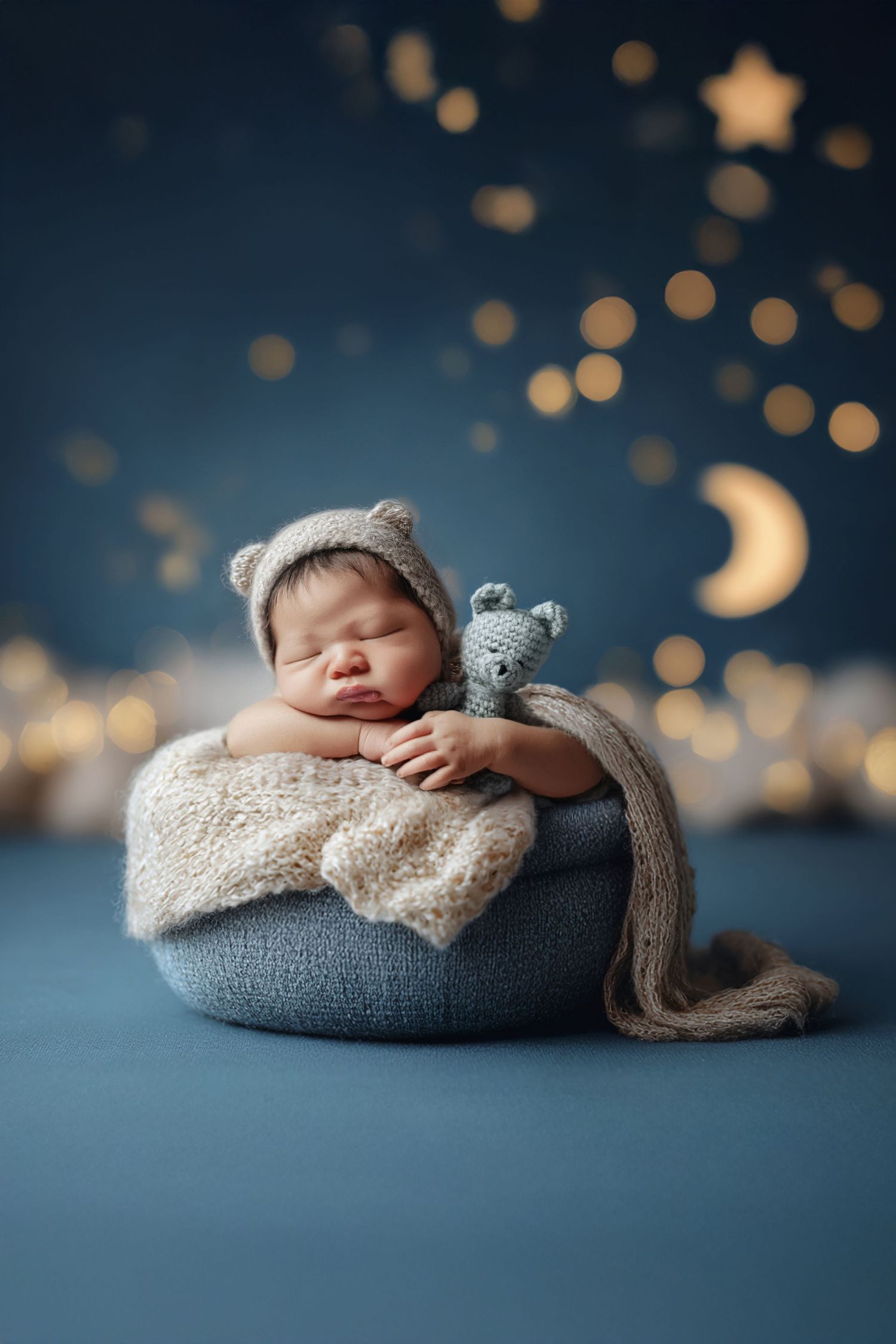 Sleeping newborn baby in bear hat holding knit toy in blue bowl with bokeh stars and crescent moon backdrop.