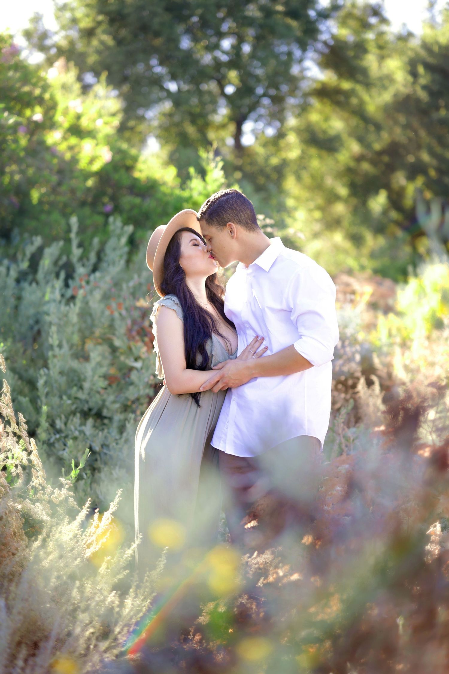 A couple shares a romantic kiss surrounded by lush greenery, woman in hat and sage dress, man in white shirt.