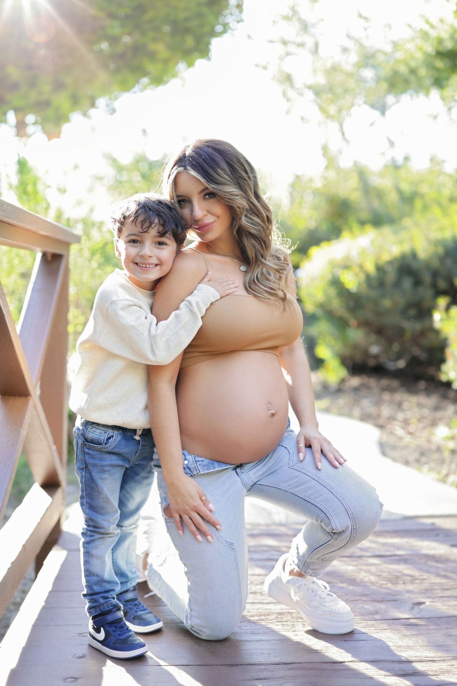Pregnant woman kneeling on wooden bridge with young boy hugging her in sunny outdoor park setting.