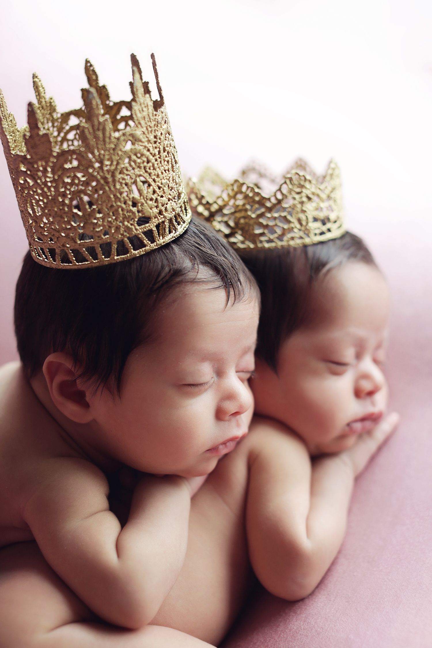 Two sleeping newborn twins wearing golden lace crowns lying together on a pink blanket.