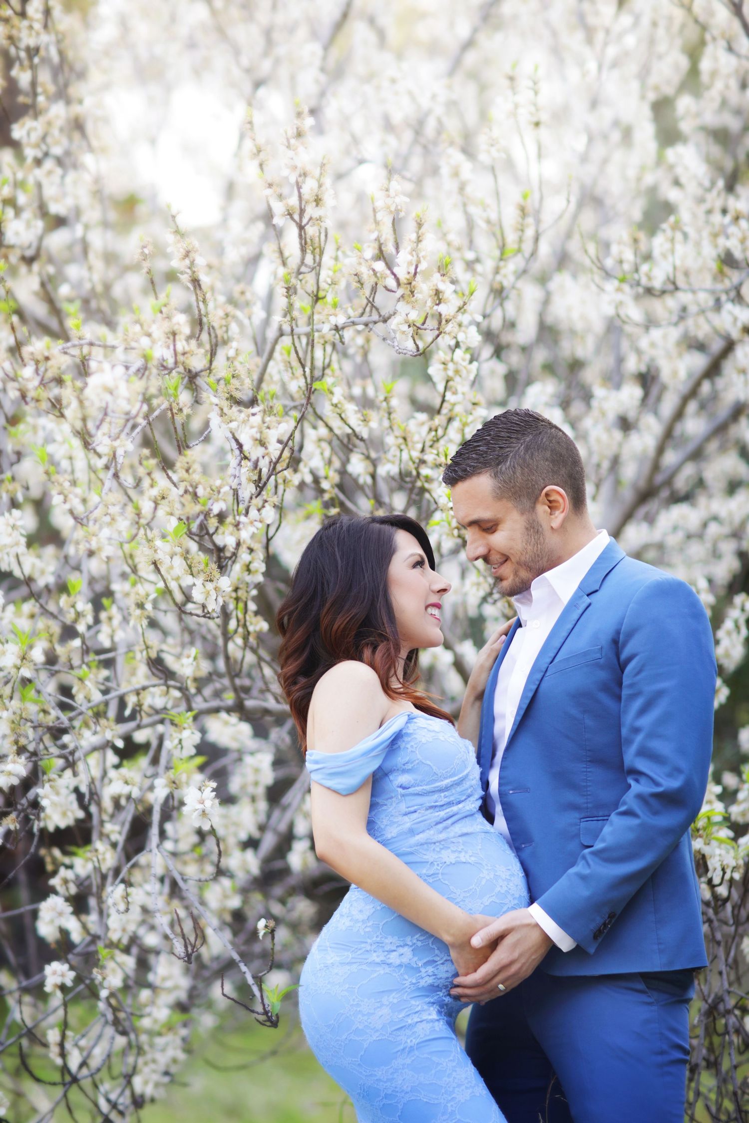A couple in matching blue outfits embraces near blooming white cherry blossom trees, smiling at each other.