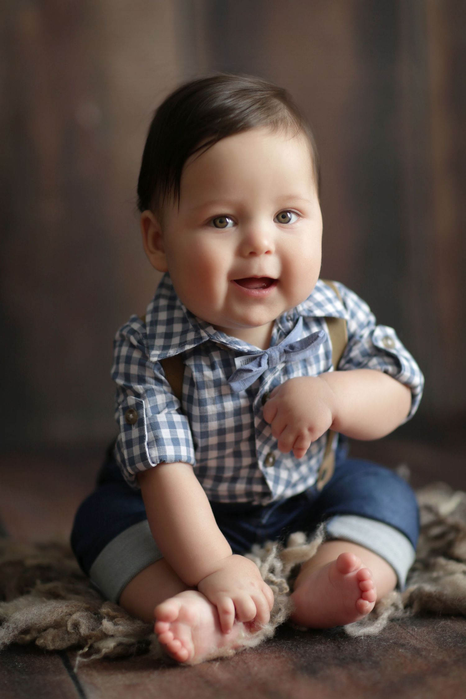 Adorable baby boy in plaid shirt and jeans sitting barefoot on rustic wooden floor, smiling happily.