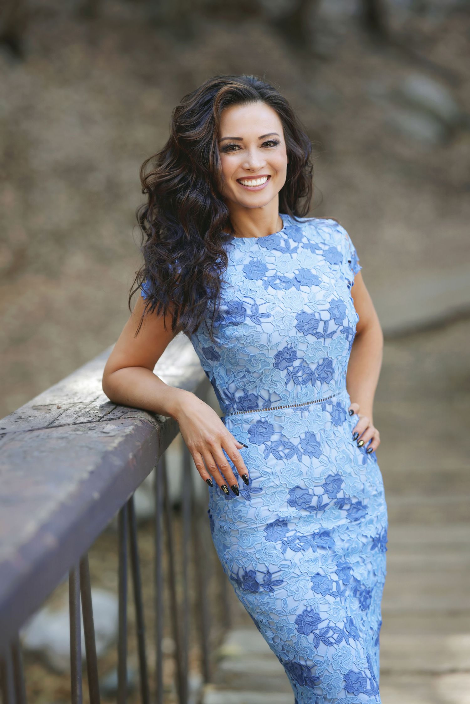 Smiling woman in blue floral lace dress leaning on railing outdoors with wavy dark hair.