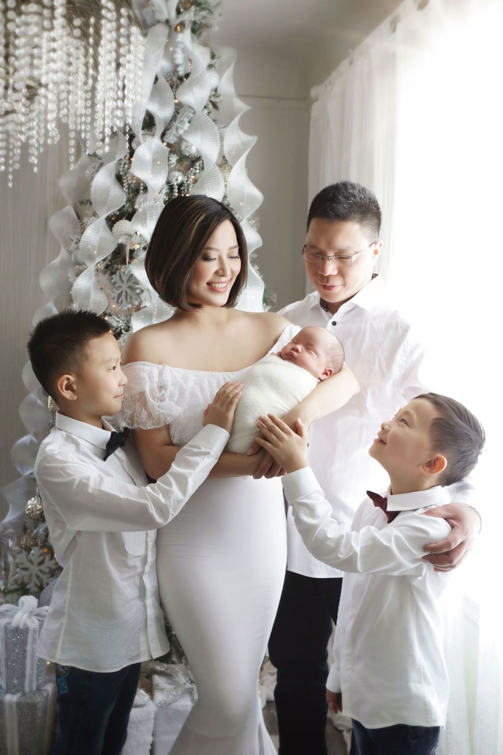 Asian family in white outfits celebrating Christmas with newborn baby near decorated holiday tree.