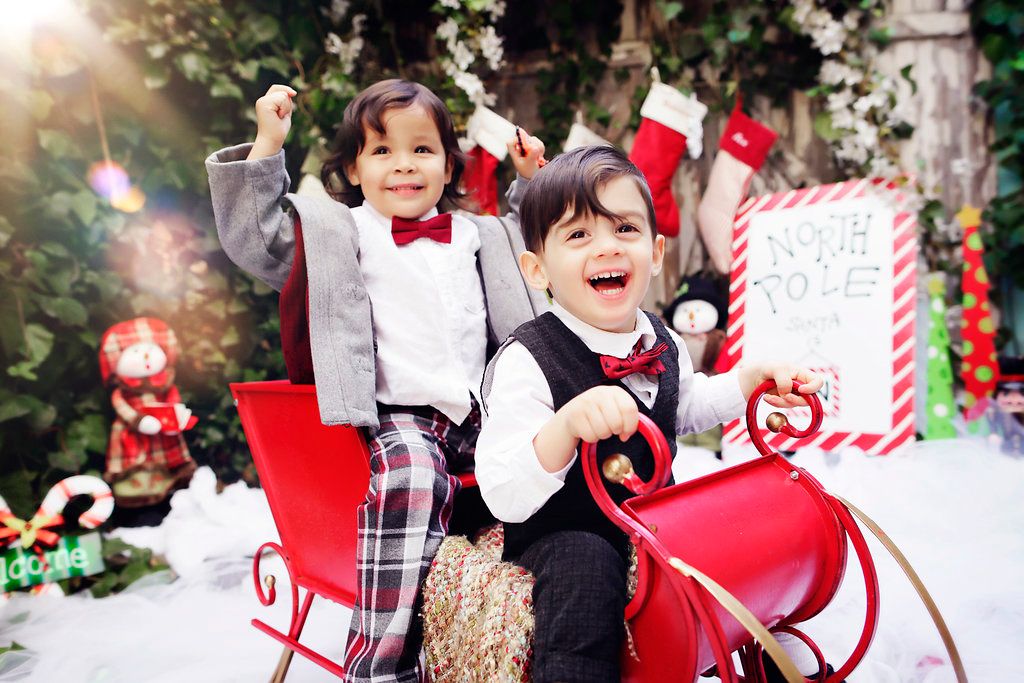 Two cheerful children in holiday outfits sit in a red sleigh surrounded by Christmas decorations and stockings.