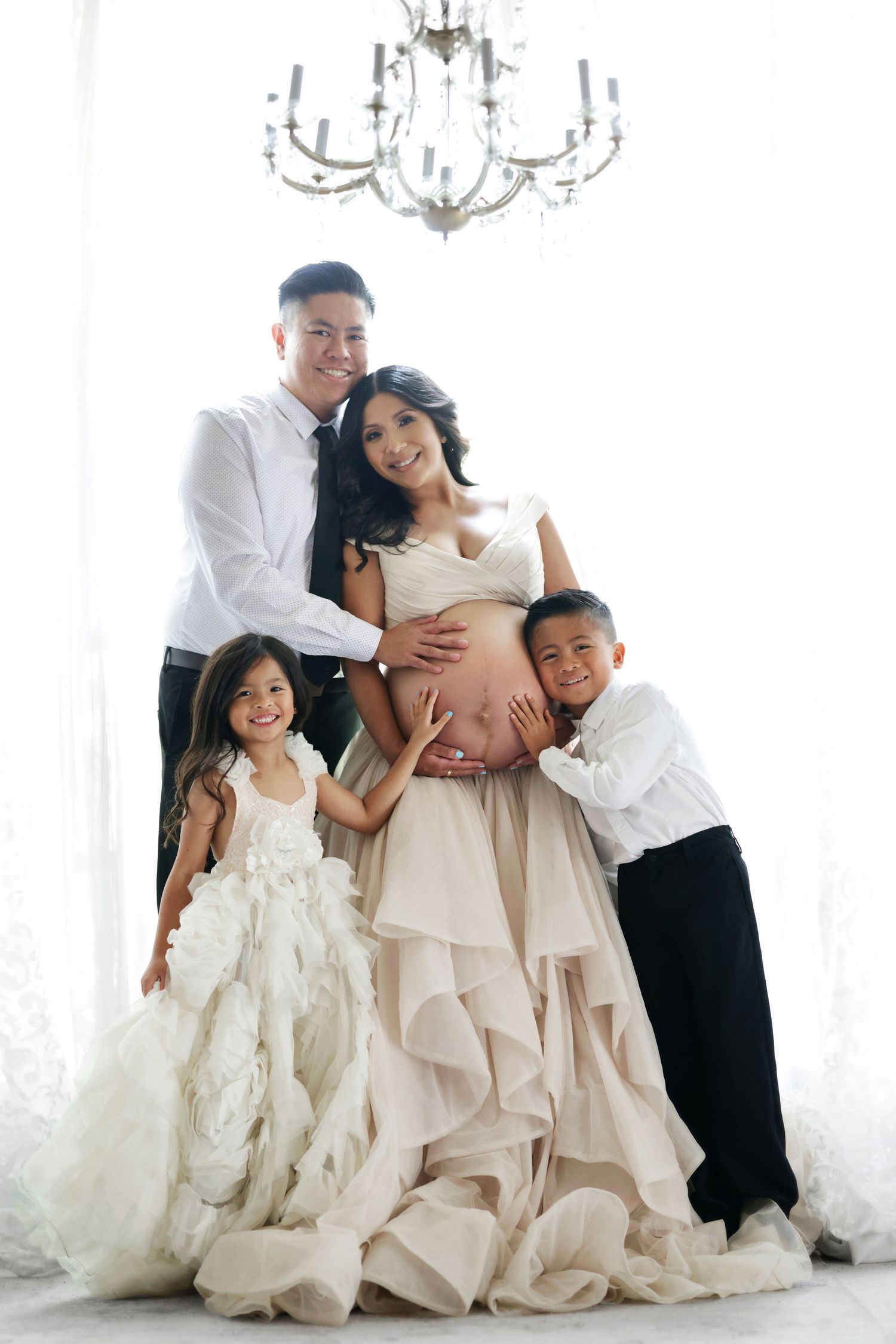 Elegant family maternity photo with pregnant mother in ruffled gown, father, and two children under chandelier.