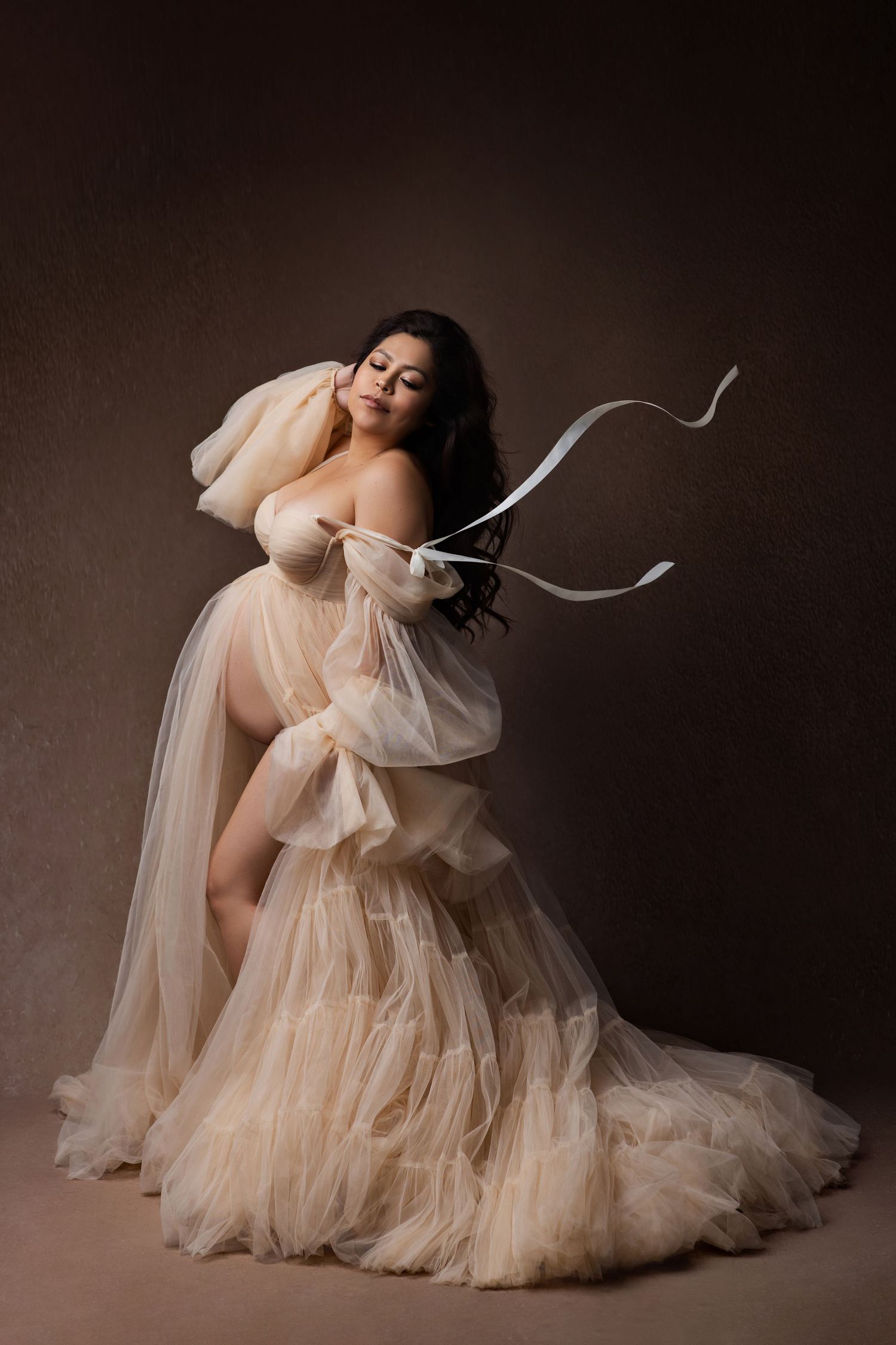 Pregnant woman in flowing cream tulle gown holding white ribbon in elegant maternity portrait against brown backdrop.