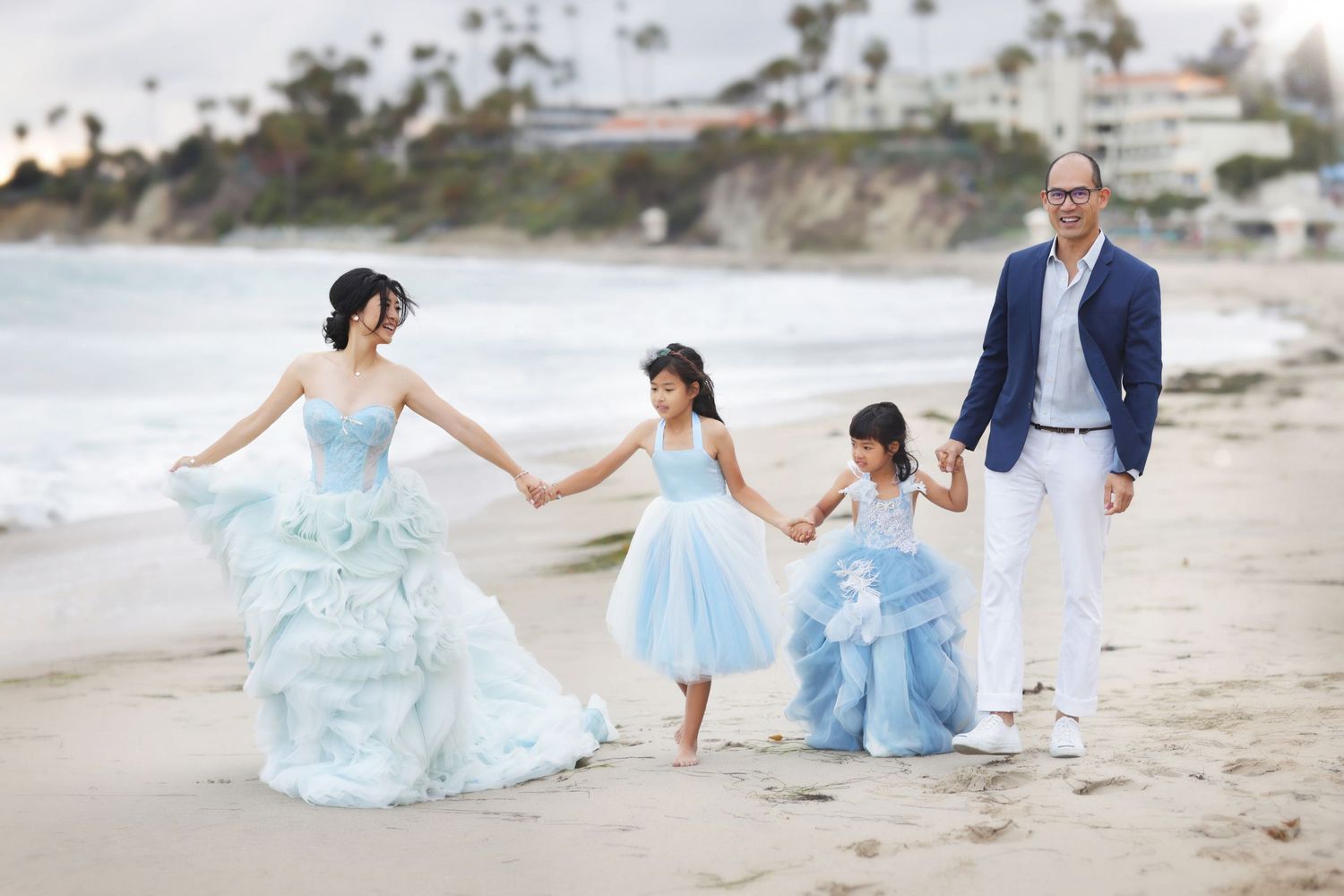 Family of four in light blue and white outfits walking hand in hand on a scenic beach.