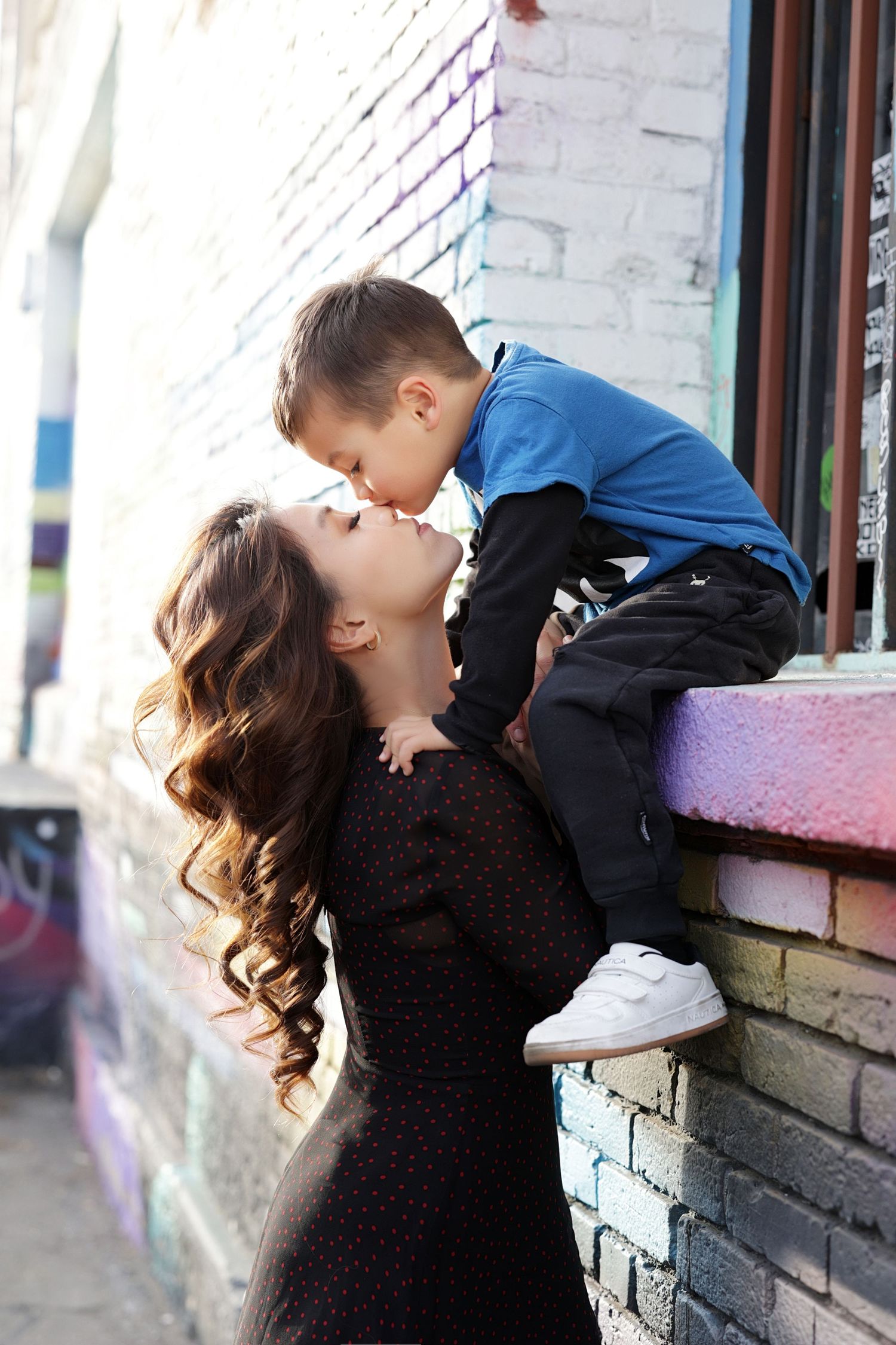 A mother and young son share a tender kiss outdoors near a colorful graffiti brick wall.