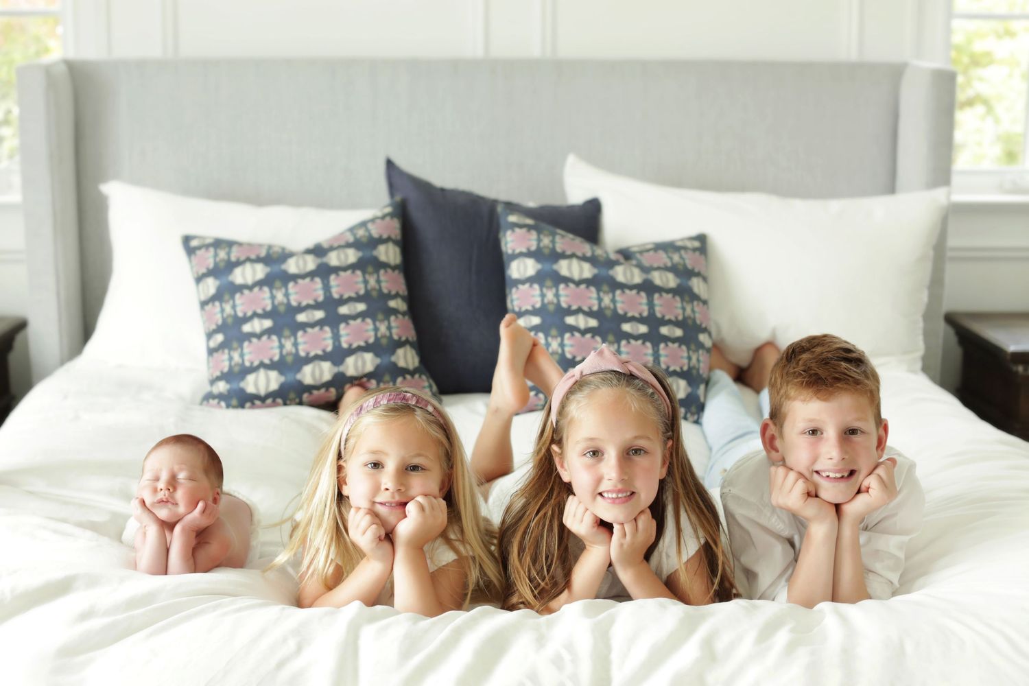 Four children lying on a white bed with decorative pillows, smiling at the camera in a bright bedroom.
