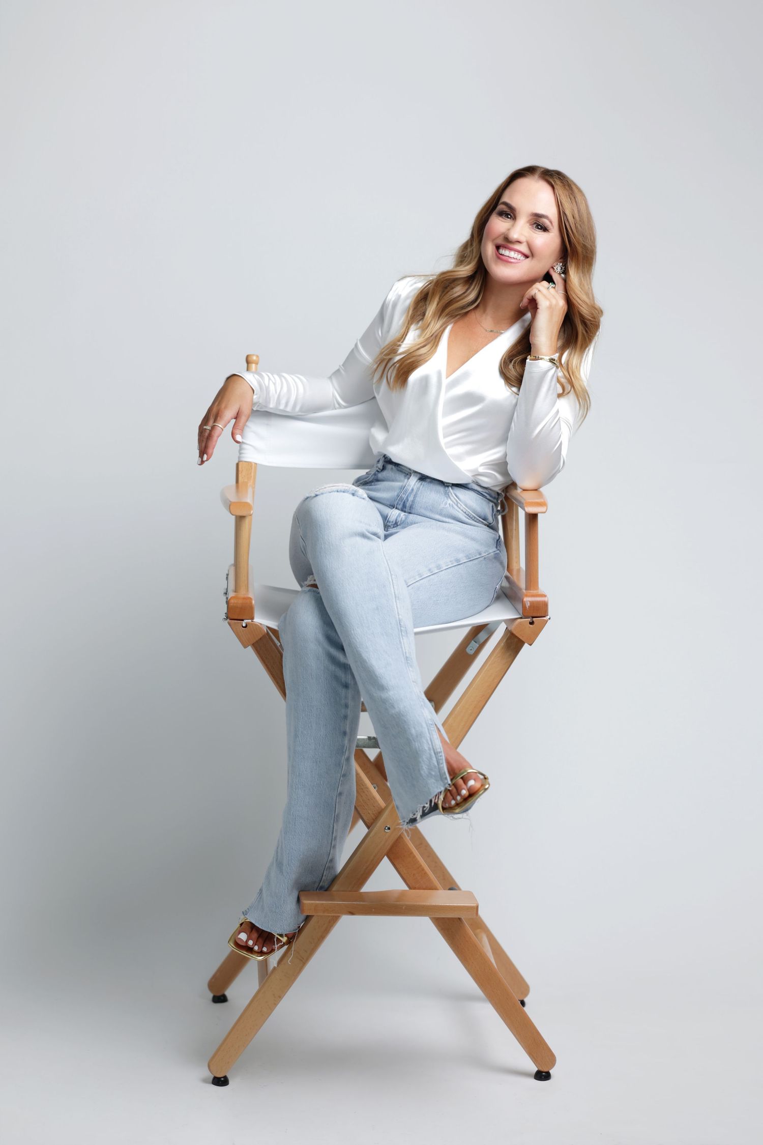 Smiling blonde woman sitting in a directors chair wearing white blouse and light blue jeans in studio.