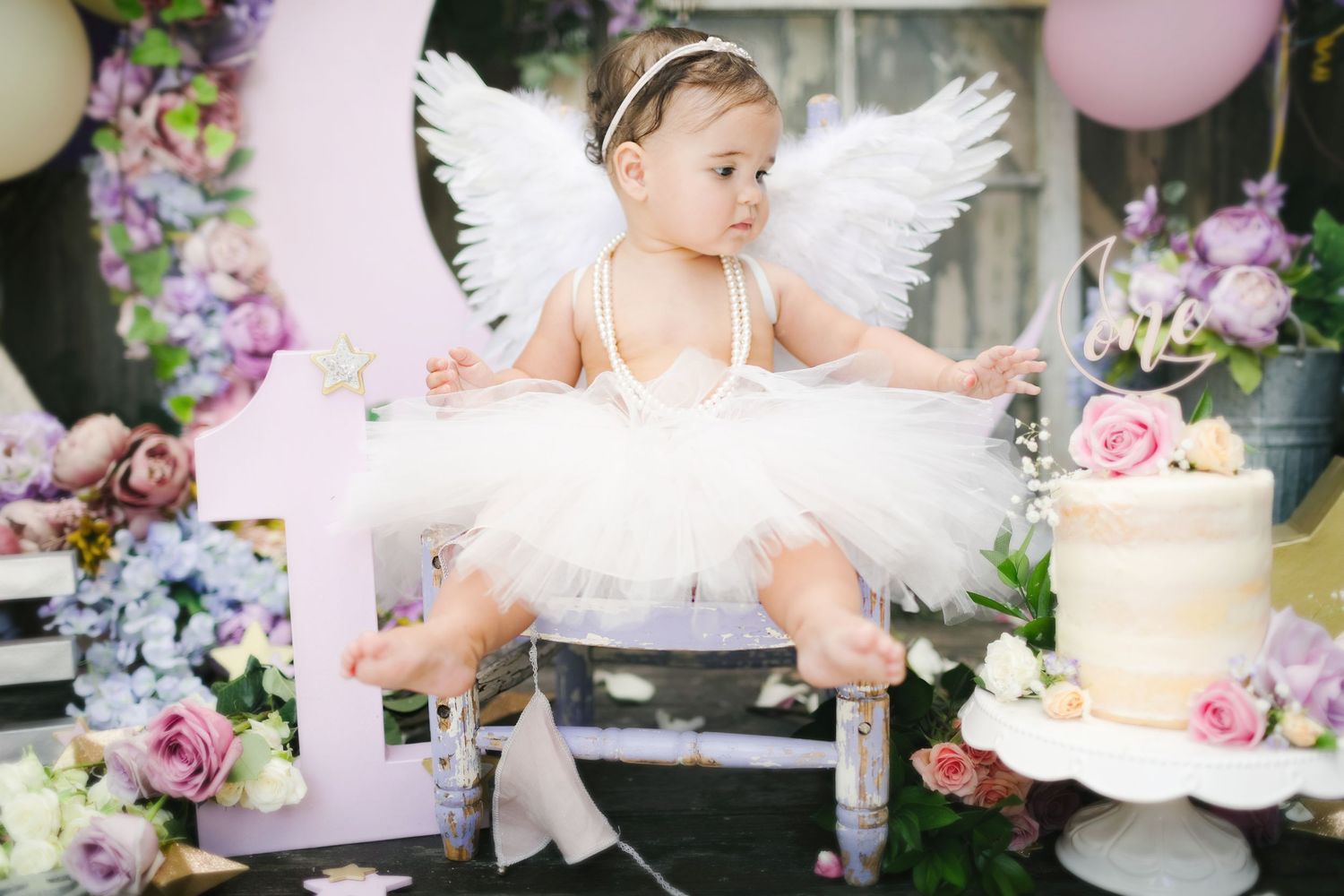 Adorable baby girl in white tutu and angel wings sits among colorful flowers for first birthday.
