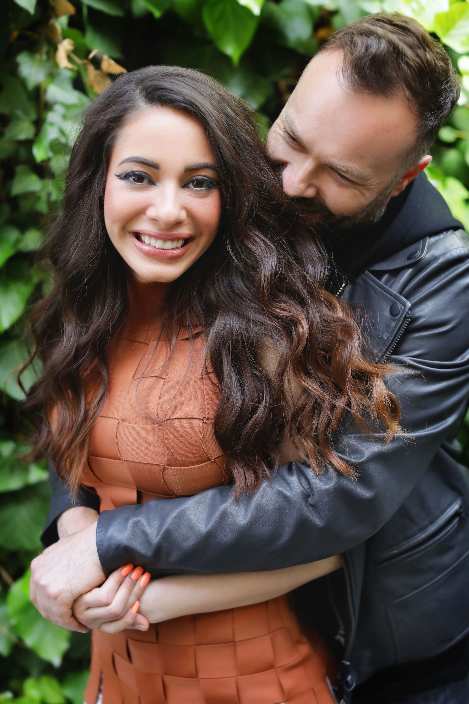 A smiling woman in an orange dress embraced by a bearded man in a black leather jacket, standing before green ivy.