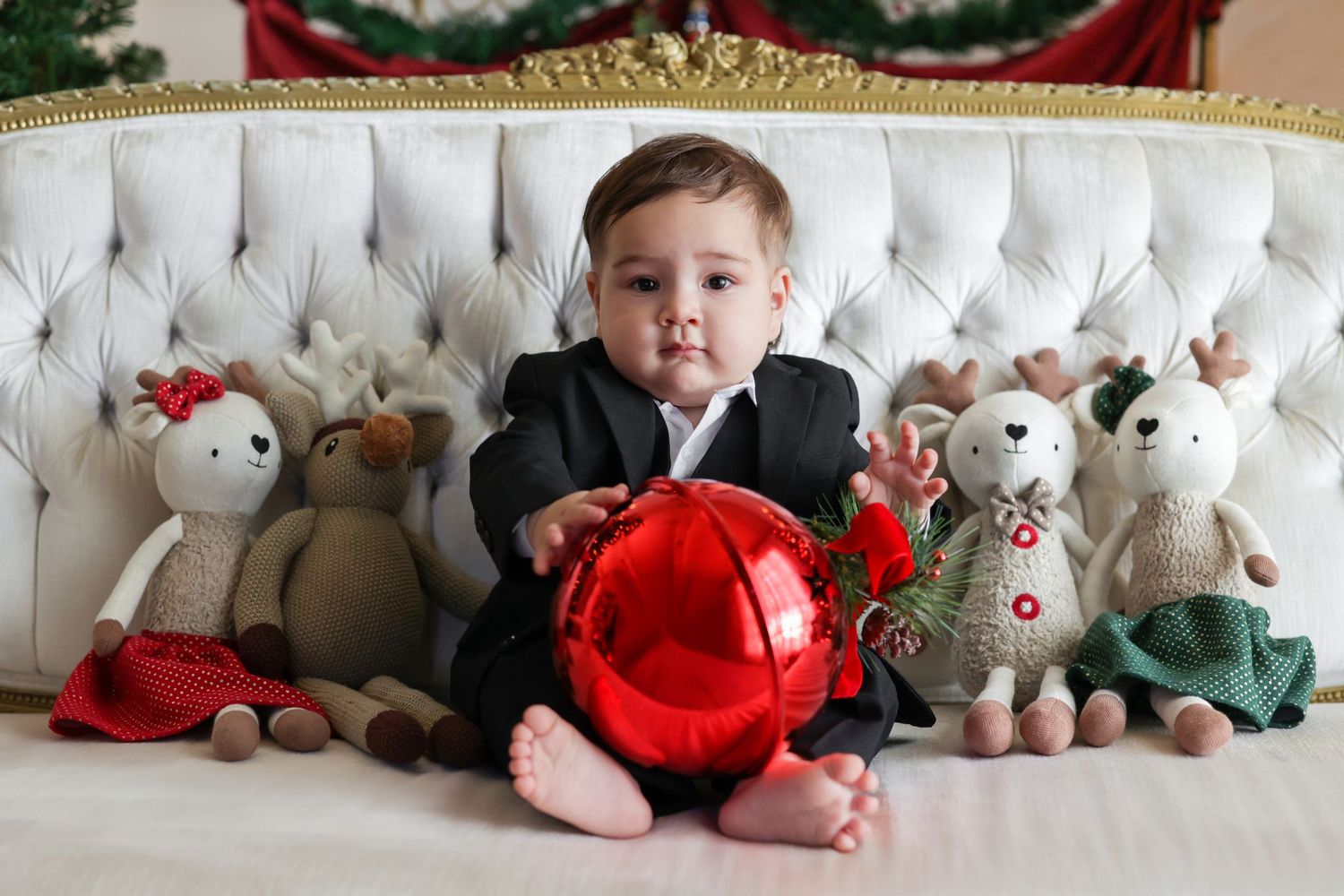 Baby in black suit holding red ornament balloon surrounded by stuffed reindeer toys on white tufted sofa at Christmas.