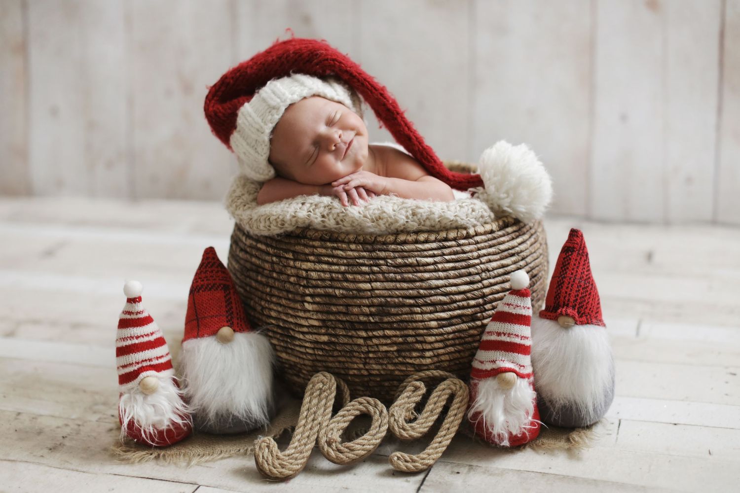 Sleeping newborn in Santa hat nestled in wicker basket surrounded by Christmas gnomes and JOY sign.