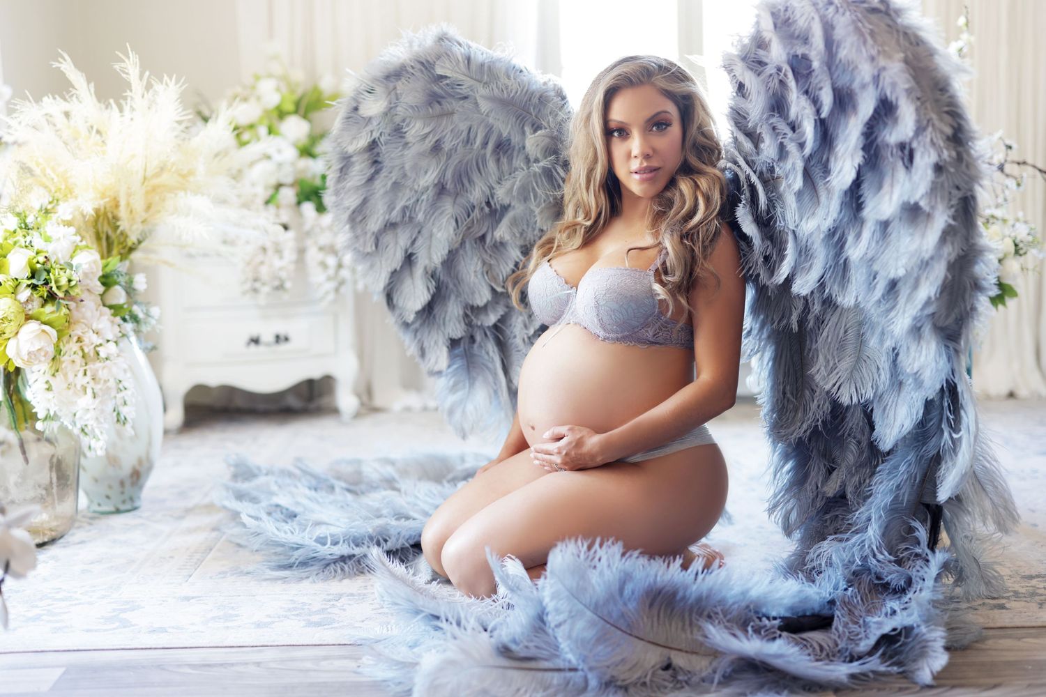 Pregnant woman in lingerie with large gray angel wings kneeling among white flowers in bright studio.
