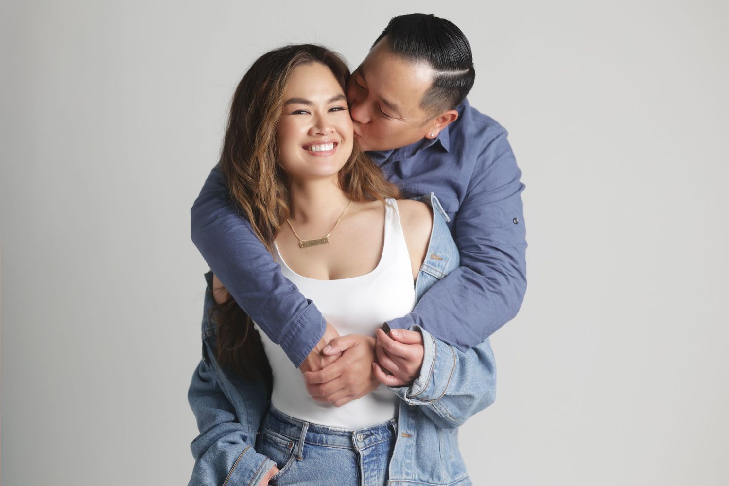 Happy couple embracing, woman smiling as man kisses her cheek, wearing casual denim outfits on gray background.