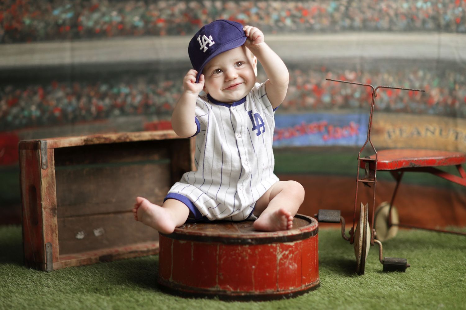 Smiling baby in LA Dodgers baseball outfit and cap sitting on vintage red drum in stadium-themed photo session.