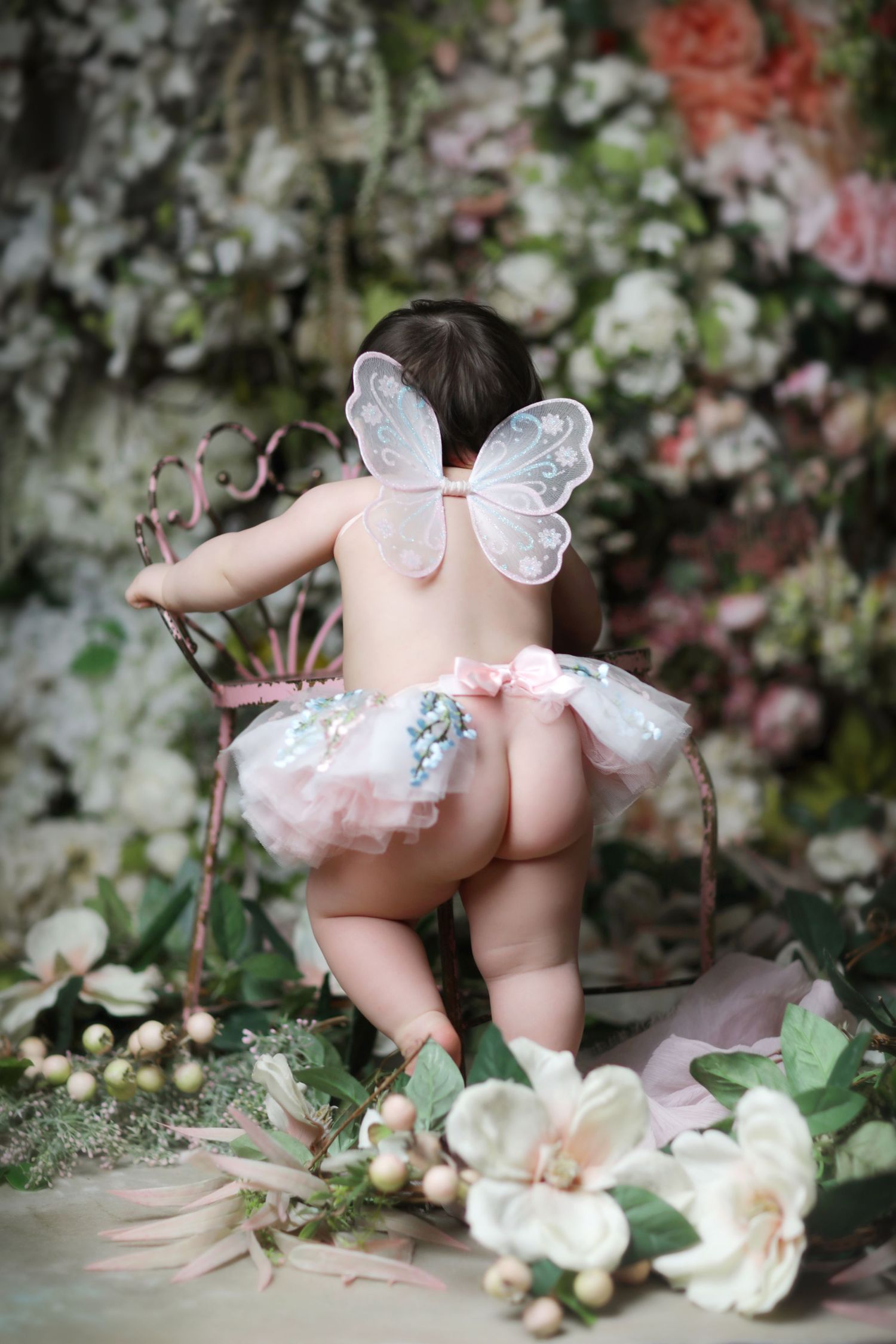 Adorable baby wearing fairy wings and pink tutu skirt standing among beautiful floral arrangement backdrop.