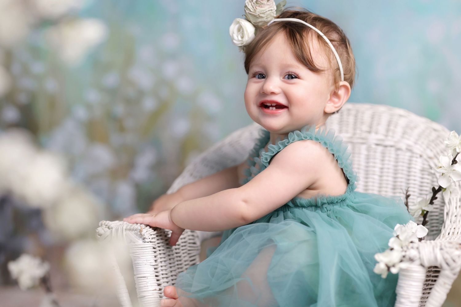 Smiling baby girl in teal tutu dress sitting in white wicker chair with floral headband.