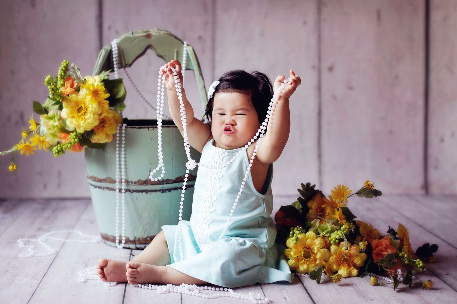 Adorable baby girl in blue dress playing with pearl necklace beside yellow flowers and vintage bucket.