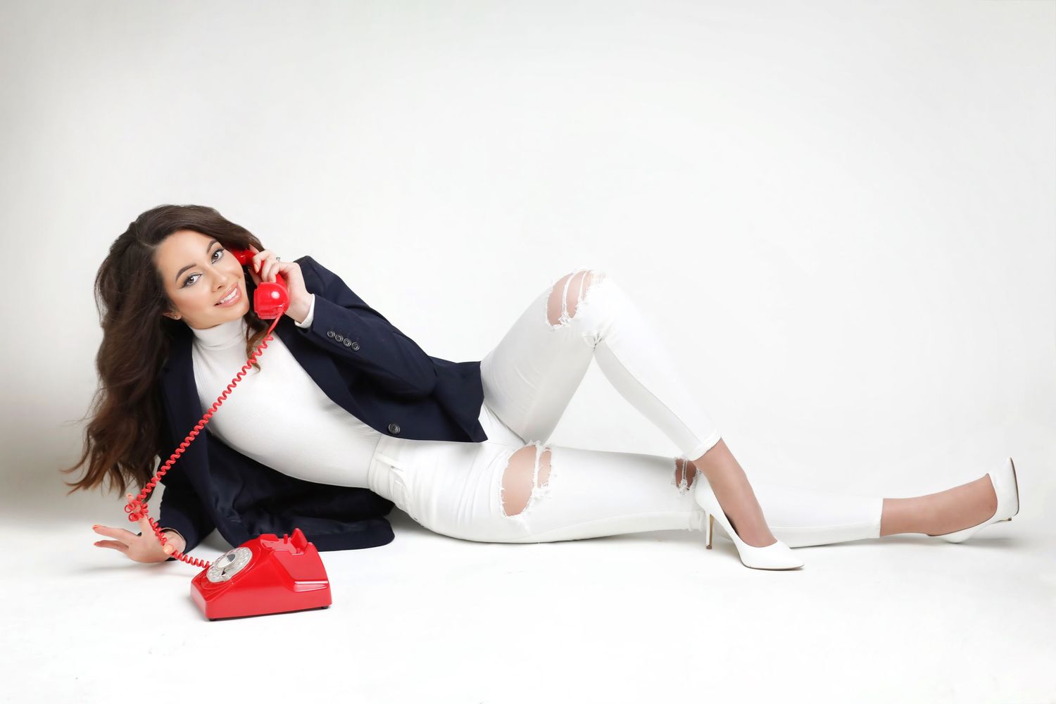 Woman in white jeans and blazer posing with red rotary telephone on white background.