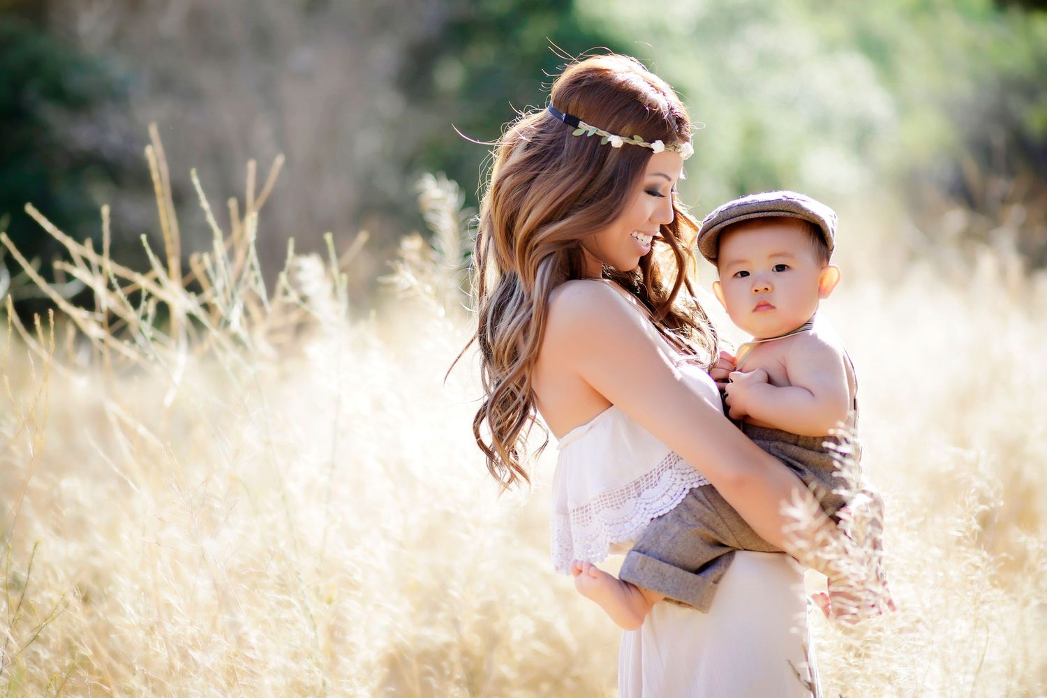Mother in white dress holding baby in hat among golden grass fields, soft sunlight bokeh background.