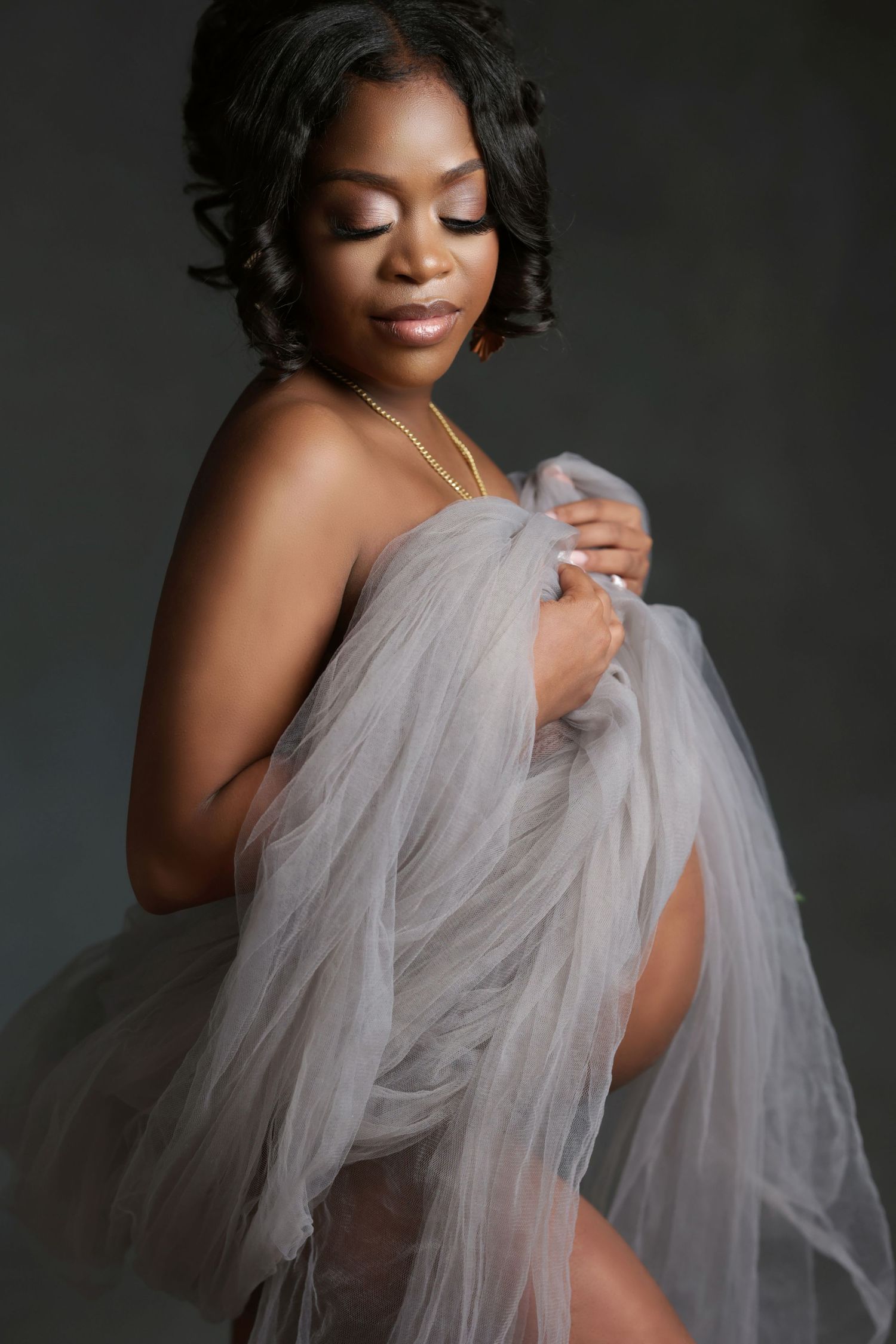 Elegant pregnant Black woman draped in sheer gray tulle fabric, posing gracefully in a studio maternity portrait.