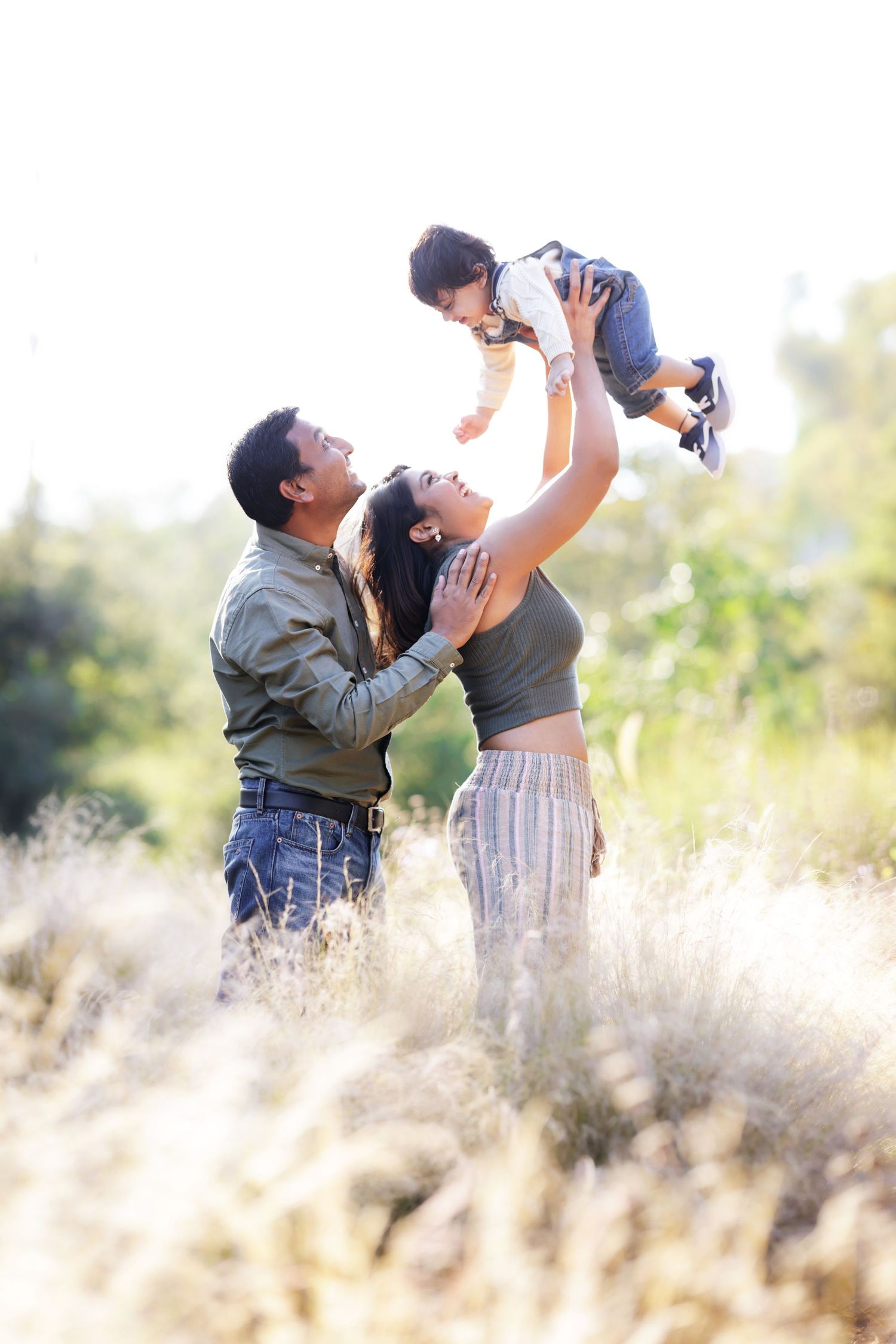 A joyful family outdoors, with a woman lifting a baby up high while a man stands beside her smiling.