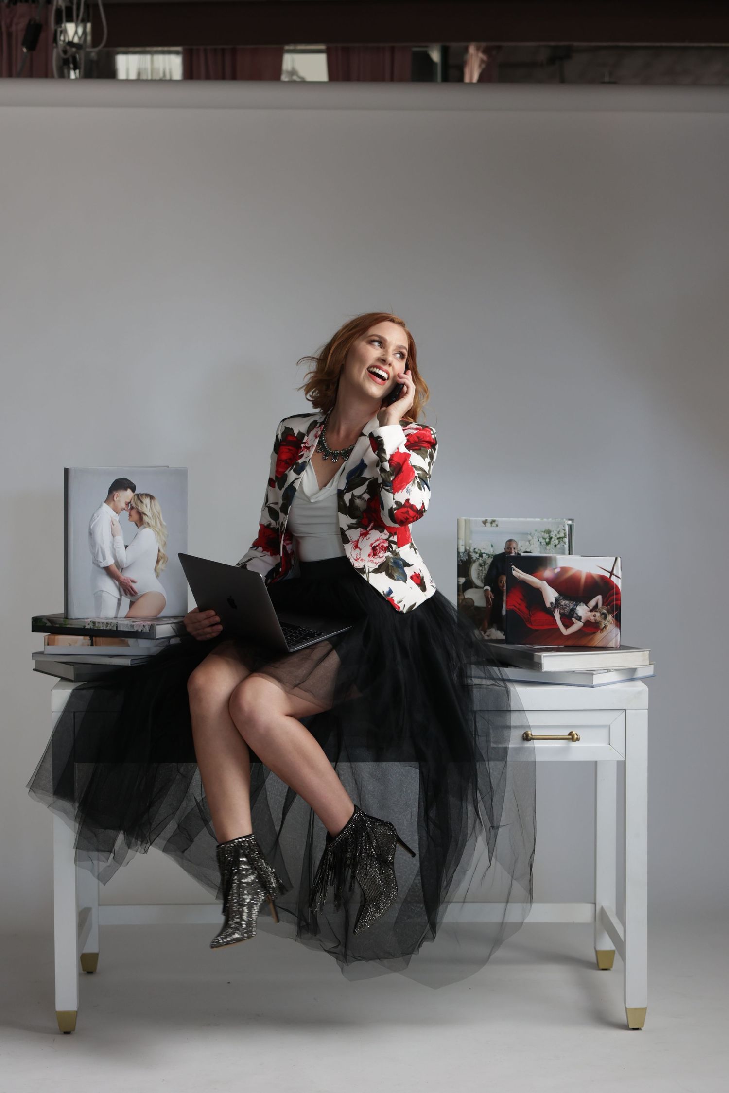 Woman in floral blazer and tulle skirt sits on desk talking on phone in studio.