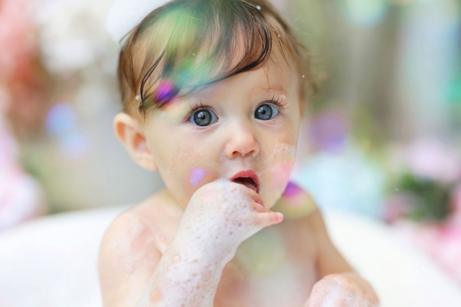 Blue-eyed baby girl with finger in mouth sits in bubble bath during first birthday celebration.