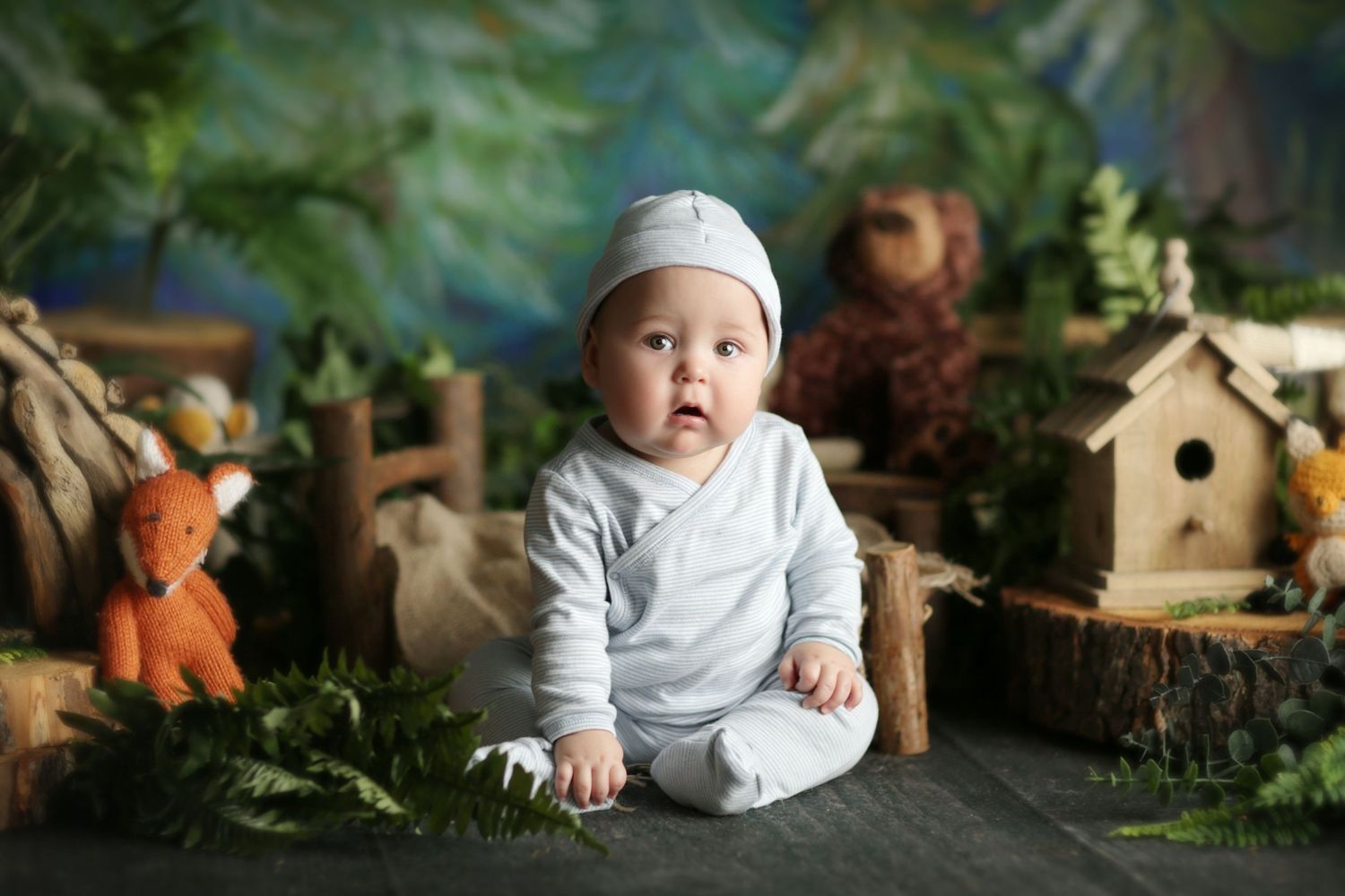 A cute baby in white outfit and hat sits among woodland decorations with ferns, birdhouse, and fox toy.