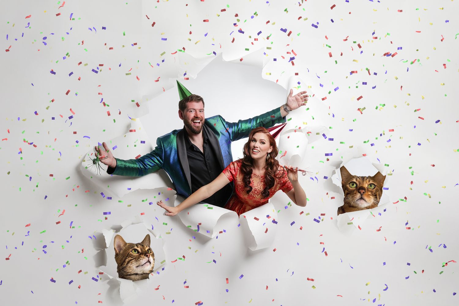 Man and woman in party attire burst through paper wall with confetti and two cats, celebrating a festive occasion.