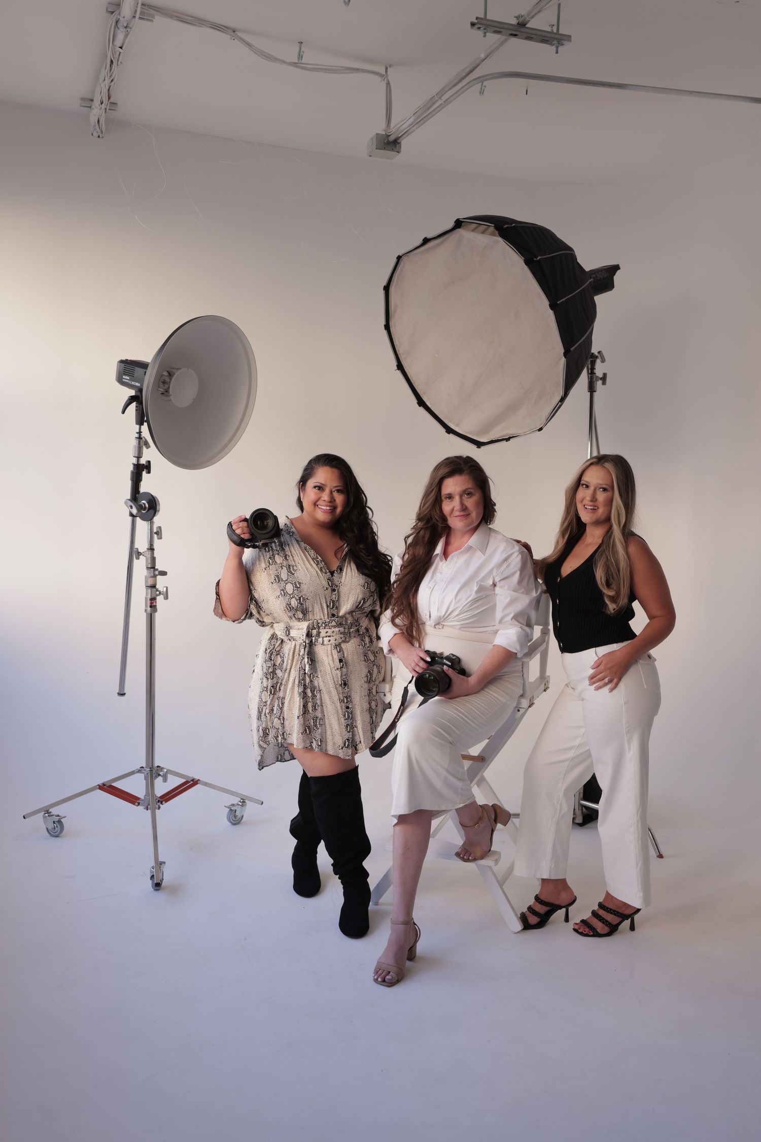 Three female photographers posing with cameras and studio lighting equipment in white studio.
