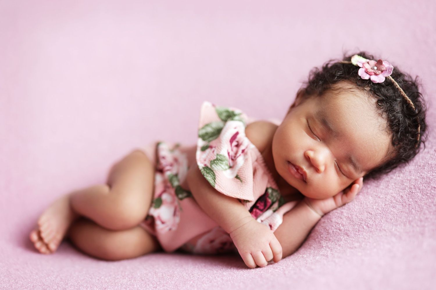 Sleeping newborn baby in floral pink romper with headband on soft pink blanket.