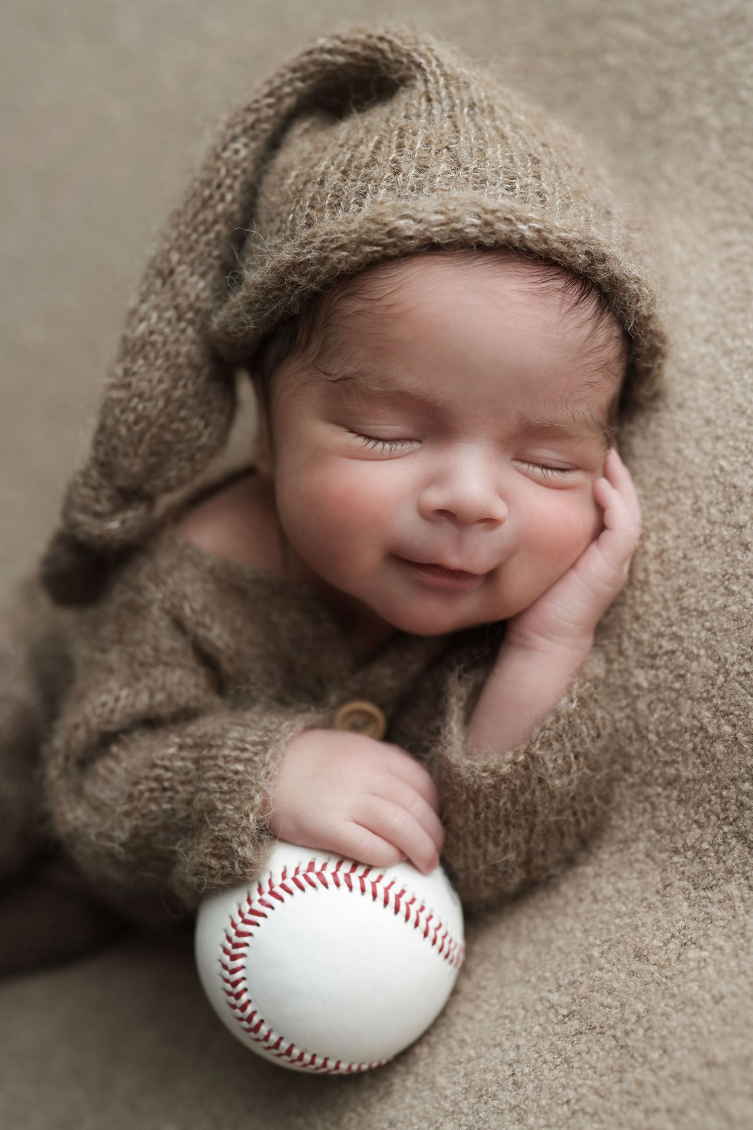 Peaceful sleeping newborn baby in brown knit hat holding baseball during professional newborn photo shoot.