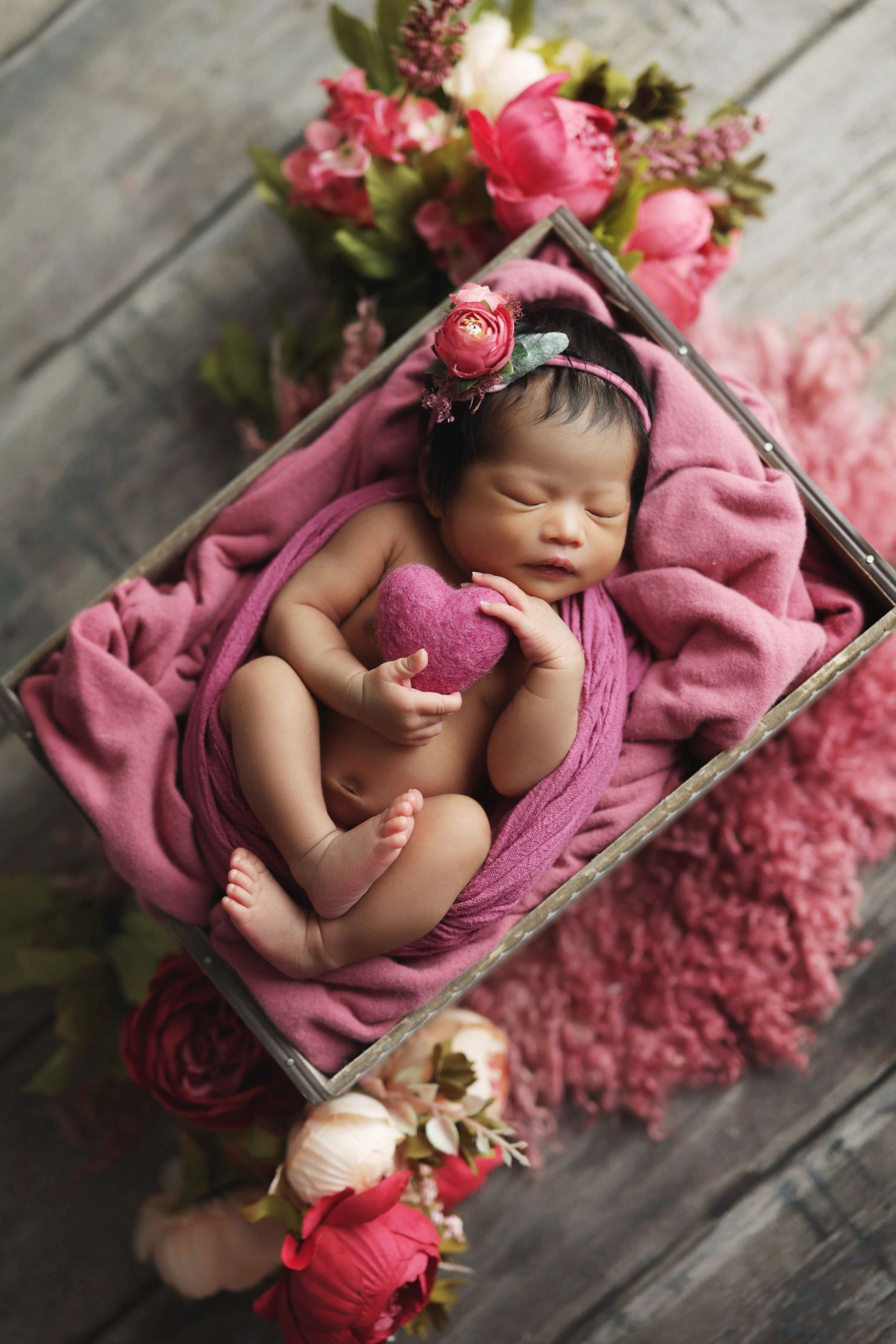 Newborn baby girl sleeping in a wooden crate with pink floral headband, wrapped in mauve cloth with flowers surrounding her.