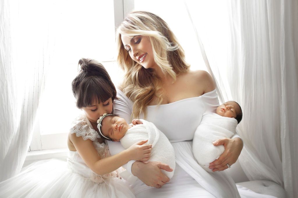 A woman in white holds a newborn while a young girl in a floral dress cuddles another baby near sheer curtains.